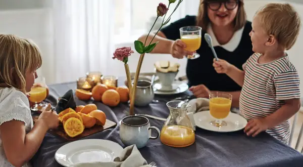 Familjen samlas kring frukostbordet med apelsinjuice och frukt. Duken är av linne, redo att stryka för nästa måltid.