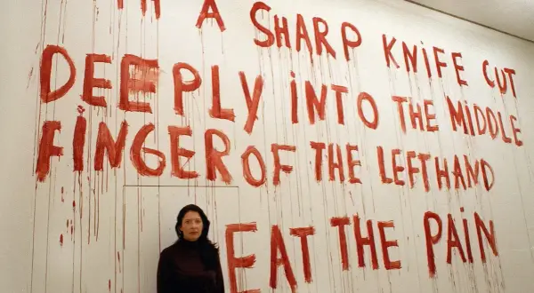 A woman stands before a wall with red, dripping text that reads "WITH A SHARP KNIFE CUT DEEPLY INTO THE MIDDLE FINGER OF THE LEFTHAND EAT THE PAIN." This is a reference to "spirit cooking.