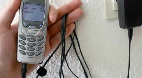 Dłoń trzyma starą Nokię, która ładuje się przez kabel. Na ekranie telefonu widnieje napis "Akku wird geladen". To klasyczny przykład technologii 3G z dawnych lat.