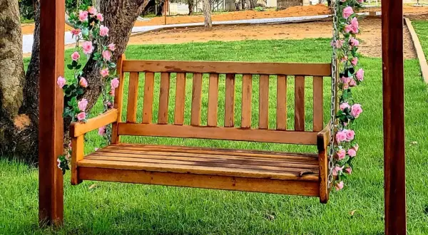 Drewniana ławka huśtawka ogrodowa ozdobiona różowymi kwiatami, na tle zielonej trawy.