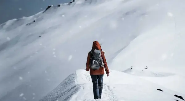Persona con zaino cammina su un crinale innevato, mentre fiocchi di neve cadono dolcemente.