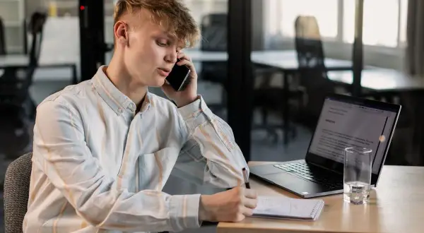 Młody mężczyzna rozmawia przez telefon, notując coś w zeszycie. Obok laptop i szklanka wody. Czy to numer 8012 co to za numer?