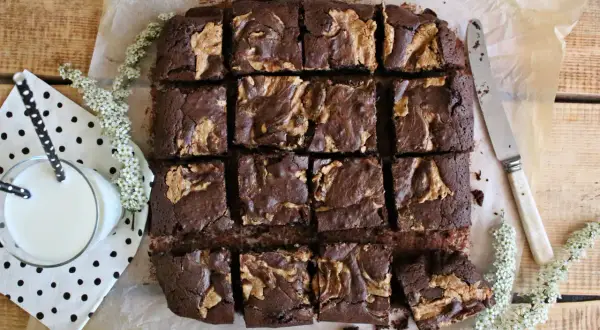 Pyszne brownie z masłem orzechowym, pokrojone w kostkę, czeka na degustację. Obok szklanka mleka i delikatne białe kwiaty.
