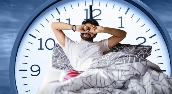 Man in bed rubs his eyes, looking tired. A large clock face behind him shows it's just past 6 AM, suggesting only six hours of sleep.