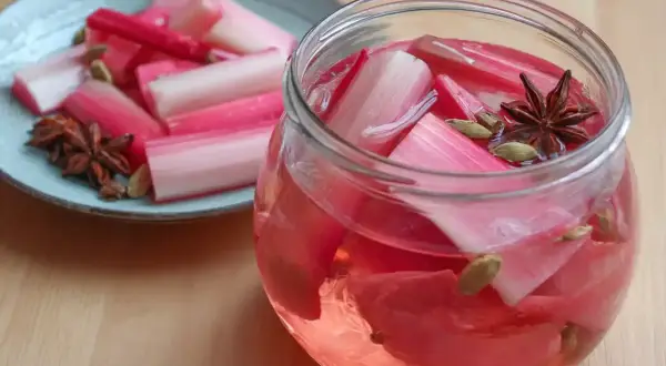 Rabarbersaft recept: färska rabarberbitar i en glasburk med kryddor som stjärnanis och kardemumma. En tallrik med rabarber och kryddor står bredvid.