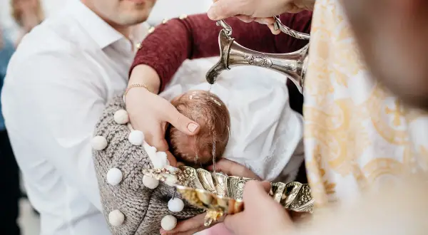 Chrzest dziecka podczas chrzcin. Woda z ozdobnego dzbanka spływa na główkę niemowlęcia.