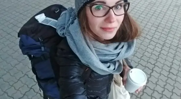 Kobieta w czapce i szaliku trzyma kubek kawy, obok plecak.