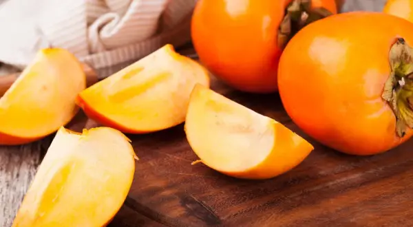 Saftige, leuchtend orangefarbene Persimmon-Früchte, einige ganz, andere in Spalten geschnitten, liegen auf einem Holzbrett.