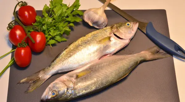 Due pesci serra freschi, pomodorini, prezzemolo, aglio e un coltello su un tagliere, pronti per una cena deliziosa.