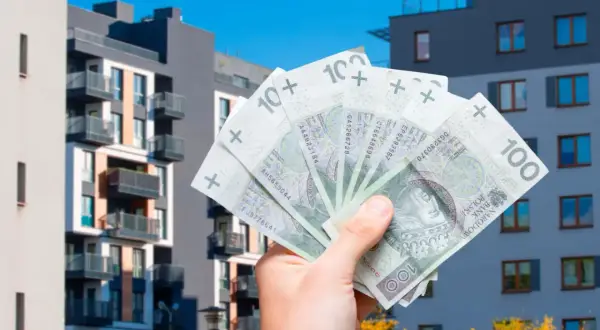 Dłoń trzyma polskie banknoty 100 zł na tle nowoczesnych bloków mieszkalnych. To symbol ulgi mieszkaniowej i możliwości zakupu własnego lokum.