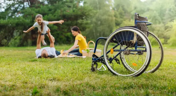 Rodzina na pikniku, obok wózek inwalidzki. Radość dziecka unoszonego przez tatę, mama obok. Wsparcie dla opiekunów: zasiłek pielęgnacyjny a świadczenie pielęgnacyjne.