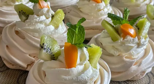 Słodkie gniazdka bezowe z bitą śmietaną, kiwi i pomarańczową dekoracją.