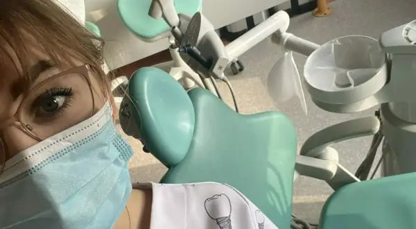 Praktyki asystentka stomatologiczna w gabinecie. Widoczny fragment twarzy w maseczce, okulary i fartuch z motywem zębów.
