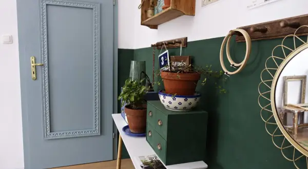 Szare drzwi pokojowe z ozdobną ramą, obok zielona komoda z roślinami. Zastanawiasz się, ile kosztuje malowanie drzwi pokojowych?