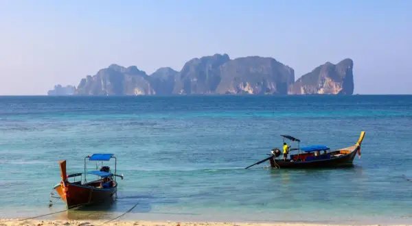 Dwa tradycyjne tajskie łodzie dryfują na turkusowej wodzie. W tle widać skaliste wyspy. Idealne miejsce na wakacje w Tajlandii, Phuket.