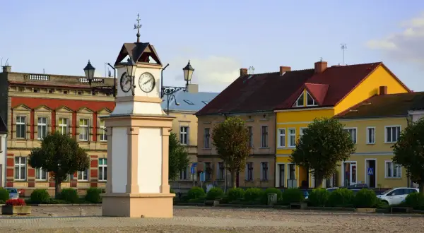 Rynek w **Lwówek Wlkp.** z zegarową wieżą po lewej i kolorowymi kamienicami w tle.