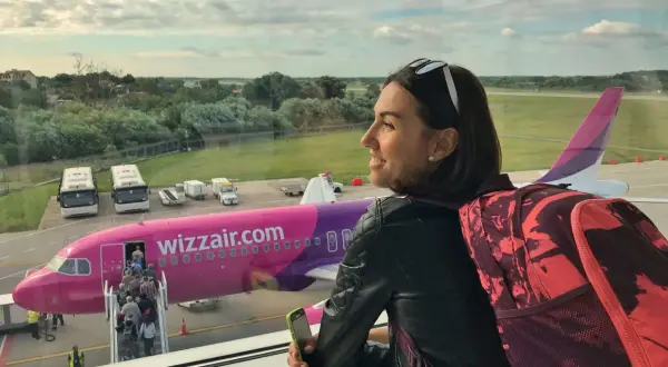 Kobieta z plecakiem i telefonem czeka na lot Wizzair. W tle różowy samolot i pasażerowie wsiadający na pokład.