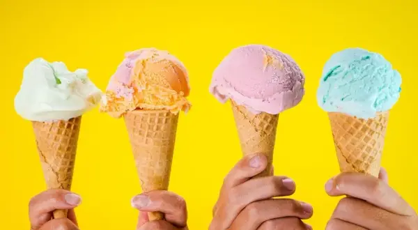 Vier Hände halten bunte Speiseeis-Sorten in Waffeln vor gelbem Hintergrund.