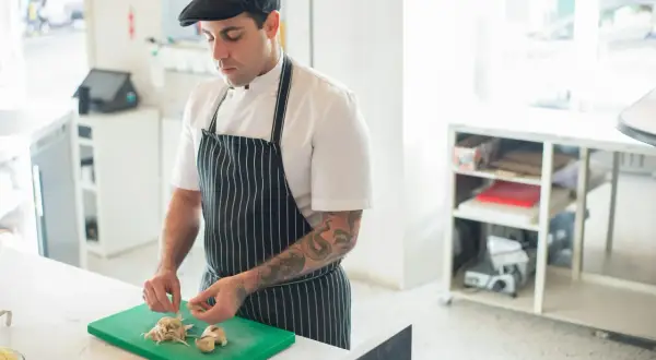 Chef con gorra y delantal a rayas, cortando champiñones en una tabla verde. Ideal para aprender a cocinar.
