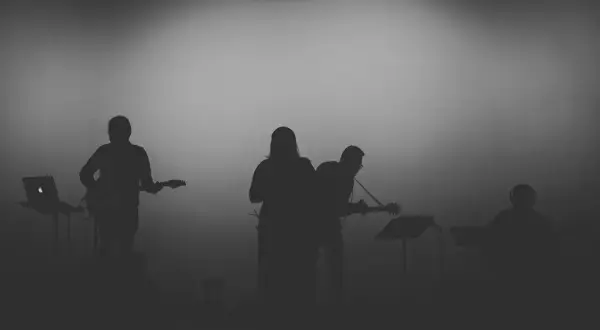 Sylwetki muzyków grających na gitarach i klawiszach na tle mglistego światła.