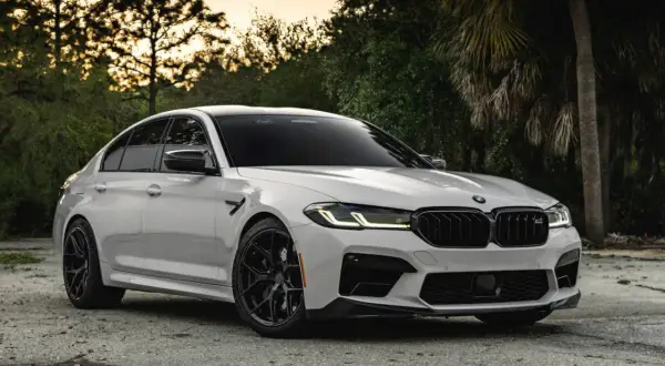 Białe BMW M5 G30 z czarnymi felgami i detalami, zaparkowane na tle drzew i zachodzącego słońca.