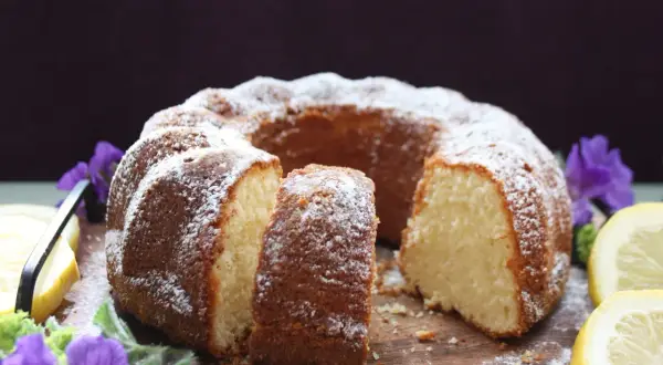En saftig citron sockerkaka, pudrad med florsocker, serveras med citronskivor och lila blommor på en träbricka.