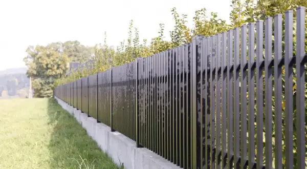 Nowoczesne, czarne ogrodzenie z pionowych profili, które wydaje się być niezgodne z granicą działki, biegnie wzdłuż betonowego murku.