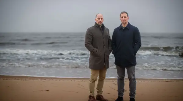 Två män står på en strand. Vågorna rullar in mot land. Det känns svårt att prata i den här blåsten.