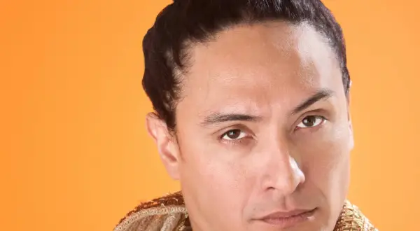 Mann mit dunklem Haar, das zu einem **Samurai-Zopf** gebunden ist, trägt ein reich verziertes Gewand vor orangefarbenem Hintergrund.