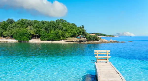 Albania - wczasy samolotem all inclusive. Drewniane molo z ławką prowadzi do krystalicznie czystej wody. Zielona wyspa w tle.
