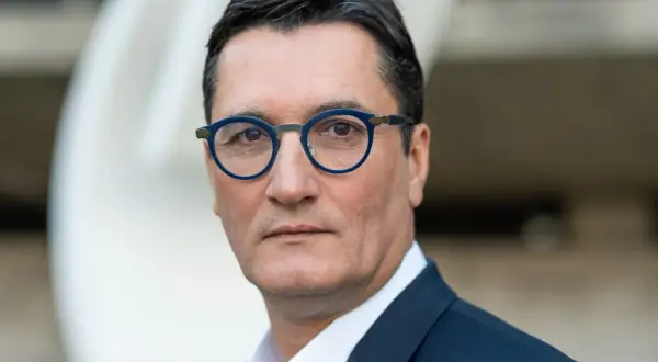 Olivier Dartigolles, un homme aux lunettes bleues, pose pour un portrait. Il est vêtu d'un costume sombre, évoquant une image de famille et de professionnalisme.