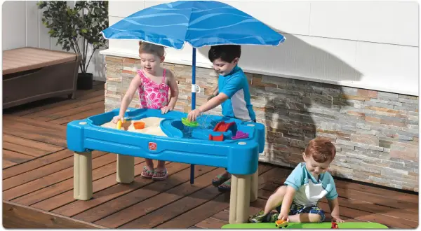 Drei Kinder genießen Sommerspiele im Sand- und Wasserspieltisch unter einem blauen Sonnenschirm.