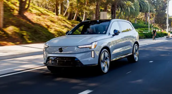 Szary Volvo EX90 jedzie po słonecznej drodze, otoczony zielenią i palmami.