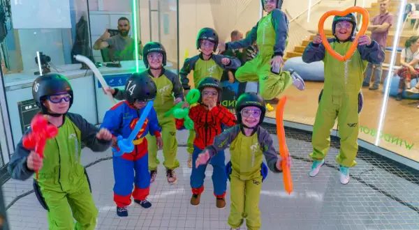 Niños con cascos y trajes verdes disfrutan de experiencias únicas, algunos con globos de colores, en un túnel de viento.