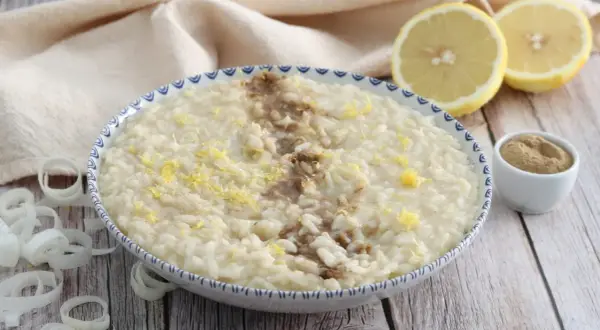 Un cremoso risotto alla liquirizia, guarnito con scorza di limone e una spolverata di polvere scura, pronto per essere gustato.