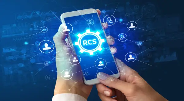 Dłonie trzymają smartfon z ikoną RCS w centrum, otoczoną siecią połączeń i symboli użytkowników na tle danych.