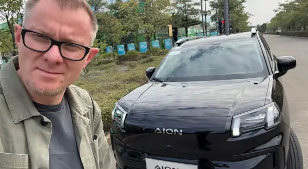 Mężczyzna w okularach pozuje do selfie obok czarnego SUV-a marki AION, jednej z nowych marek samochodów.
