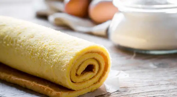 Rotolo di pasta biscotto per torte a strati, pronto per essere farcito. Sullo sfondo, uova e zucchero.