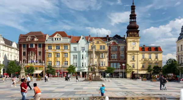 Kolorowy plac w Ostrawie z ludźmi i zabytkowymi budynkami w tle.