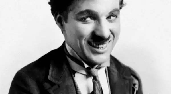 Charlie Chaplin w meloniku i garniturze, z uśmiechem, jakby opowiadał dowcip swoim dzieciom.