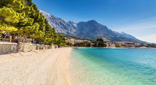 En vacker strand med turkost vatten och en bergskedja i bakgrunden. En promenadstråk kantas av träd och stenmurar, perfekt för en avkopplande stund vid makarska strandpromenad.