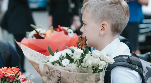 Chłopiec w białej koszuli trzyma bukiet białych kwiatów, na tle innych bukietów i ludzi.