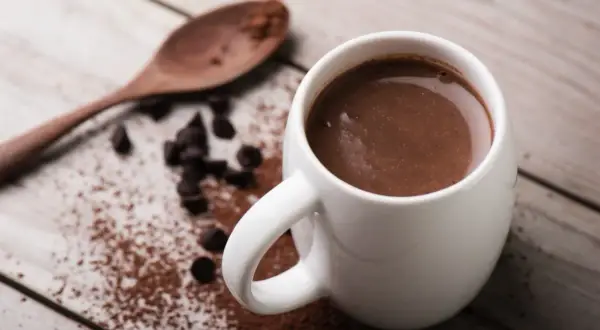 Tazza di cioccolata calda con cacao amaro, gocce di cioccolato e un cucchiaio di legno su un tavolo di legno.