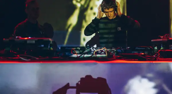 DJ w słuchawkach przy konsoli Pioneer DJ, w tle inny DJ i projekcja.