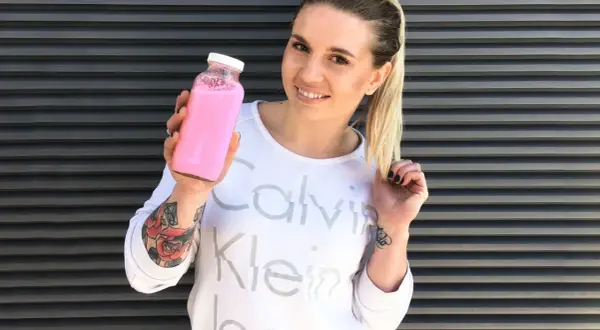 Uśmiechnięta kobieta z tatuażami trzyma różowy shake proteinowy zamiast posiłku.