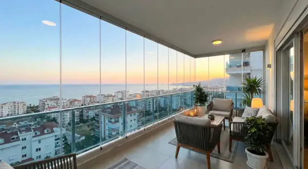 Terraza moderna con cortinas de cristal, muebles de exterior y vistas al mar al atardecer.