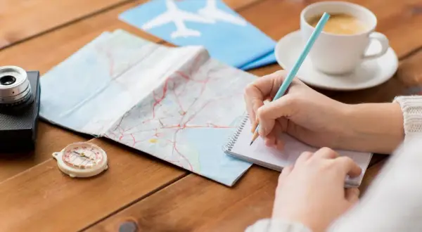 Ręce piszą listę rzeczy na wyjazd za granicę. Obok mapa, kompas, aparat i kawa.