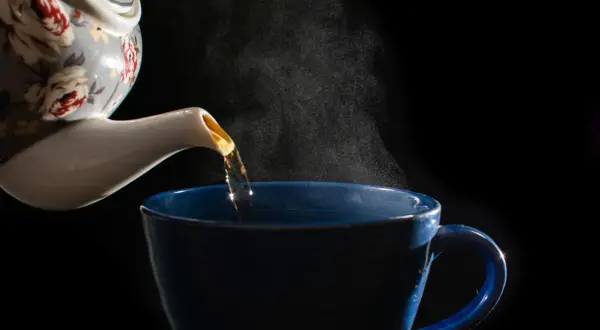 Heißer Estragon Tee wird aus einer blumengeschmückten Kanne in eine blaue Tasse gegossen. Dampf steigt auf.