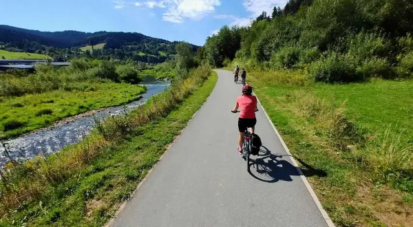 Rowerzyści przemierzają malowniczą trasę velo Krynica wzdłuż rzeki, otoczeni zielonymi wzgórzami i lasami.