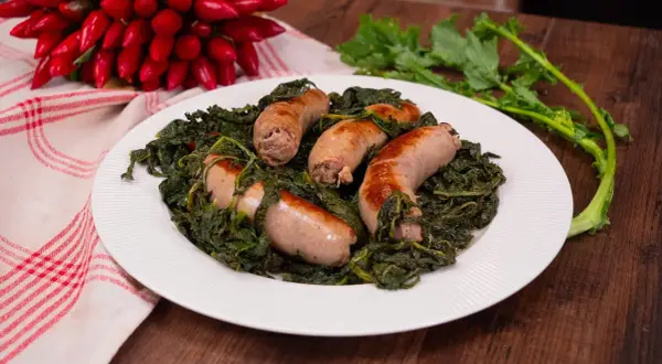 Salsiccia e friarielli, un piatto tradizionale napoletano, servito su un piatto bianco con peperoncini rossi sullo sfondo.